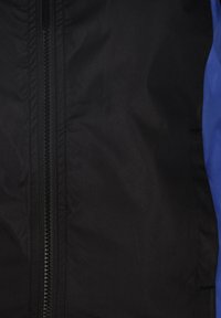 Tessuto nero con una texture liscia presenta una cerniera centrale e un accento blu sul lato destro. Progettato per l'abbigliamento esterno, materiale resistente.