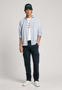 Lichtblauw gestreept overhemd over een wit T-shirt met bedrukte tekst, marineblauwe cargobroek en witte sneakers. Een donkergroen baseballpet wordt gedragen.