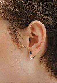 Pendiente de aro plateado con una gema azul y pequeños acentos claros. Vista en primer plano del pendiente usado en la oreja, mostrando su diseño y textura.