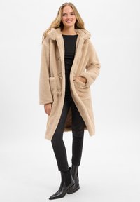 Frau in einem beigen, kapuzenartigen Teddy-Mantel, schwarzem Oberteil, schwarzen Skinny-Jeans und schwarzen, hüfthohen Stiefeln mit Absatz, die vor einem einfarbigen Hintergrund steht.