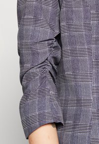 Chemise à carreaux gris présentant un mélange de grands et petits carreaux avec des accents blancs, fabriquée en tissu texturé. Manche froncée au niveau du poignet.