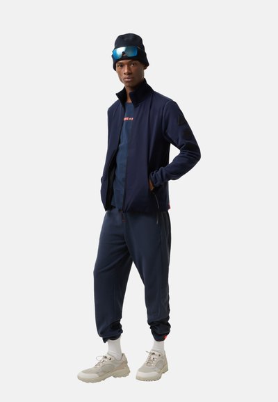 Marine sportoutfit met een ritsjack met hoge kraag en textuuraccenten, gecombineerd met bijpassende joggers en lichte sneakers.