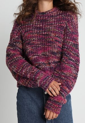 Žena v barevném fialovo-růžovém hrubě pleteném svetru s dlouhými rukávy, v kombinaci s modrou džínovou sukní, s rukama překříženýma v pase.