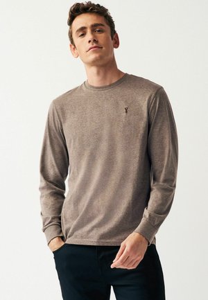 Langærmet crew-neck skjorte i lysebrunt stof med subtil tekstur. Lille logo på brystet, kombineret med sorte bukser.