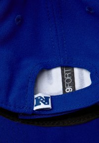 Gorra azul con forma estructurada, que presenta un visor negro, una etiqueta de tela con la inscripción "9FORTY" y un logo con estrellas sobre un fondo blanco.