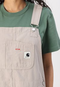 Beige overall met verstelbare bandjes, met een voorkeurszak en een klein zwart-wit logo. Gelaagd over een groene t-shirt.