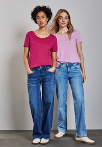 Dos mujeres posan con ropa casual, luciendo una camiseta de manga corta magenta y otra camiseta de manga corta rosa claro, acompañadas de jeans de pierna ancha.