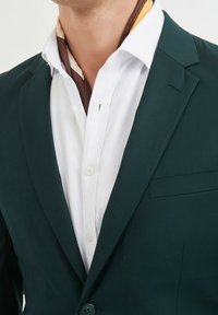 Veste vert sombre avec revers crantés sur une chemise blanche à boutons. Une écharpe marron et jaune est visible au niveau du cou. Texture subtile dans le tissu.