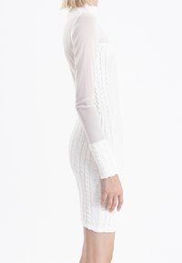 Robe blanche bodycon avec un design en maille texturée, manches longues en voile transparent et une silhouette ajustée, créant un profil épuré.