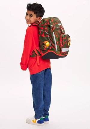 Junger Junge lächelt, trägt einen roten Pullover, blaue Jeans, weiße Turnschuhe und einen großen grünen und roten Rucksack mit Muster.