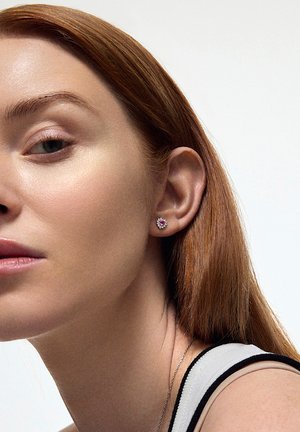 Srebrne kolczyki wkręty z różowym kamieniem w centrum, otoczone grupą mniejszych, przezroczystych kamieni, noszone na uchu modelki z czerwonymi włosami.