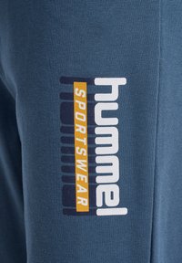 Zilas sporta bikses, izgatavotas no mīksta auduma, ar baltu "hummel" logotipu un "SPORTSWEAR" dzeltenā krāsā sānos, papildinātas ar svītrām.
