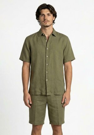 Uomo che indossa una camicia verde oliva a maniche corte con bottoni e pantaloncini abbinati, in piedi davanti a uno sfondo bianco semplice.