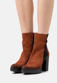 Bottines en daim marron avec un panneau latéral noir, talon haut carré et fermeture éclair latérale. Finition texturée avec un bout arrondi.