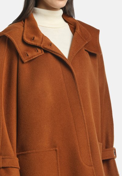 Donna che indossa un cappotto di lana marrone sopra un maglione a collo alto crema, che mostra i dettagli del collo, delle tasche e delle cinghie sulle maniche.