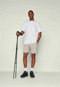 Άνδρας φορεί λευκό μπλουζάκι και μπεζ σορτς, στέκοντας με το ένα χέρι στην τσέπη και κρατώντας μαύρα σακίδια πεζοπορίας με το άλλο χέρι.