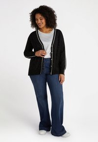 Cardigan noir avec des bordures blanches et des détails scintillants, porté sur un haut clair, associé à un jean évasé bleu et des chaussures blanches.