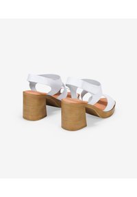 Sandalias de tacón bajo de cuero blanco con tiras anchas, parte trasera abierta y un tacón texturizado de color marrón claro. Plantilla de color rosa suave visible.