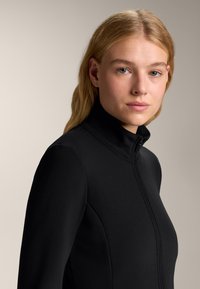 Czarna zapinana na zamek kurtka z wysokim kołnierzem, miękką fakturą i dopasowanym krojem. Szczegóły szycia podkreślają smukły wygląd.