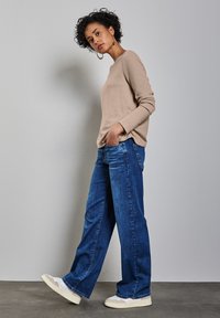 Beige stickad tröja, blå vida jeans och beige sneakers. Lockigt hår, händerna i fickorna, stående mot en ljusgrå vägg.