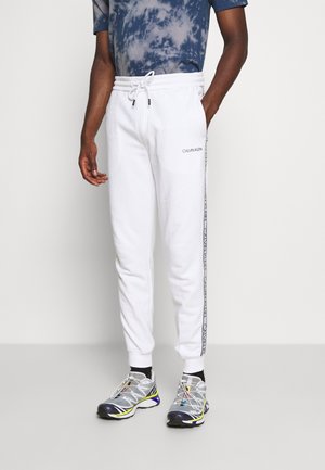 Mężczyzna noszący białe spodnie dresowe Calvin Klein z bocznym logo oraz szaro-żółto-niebieskie buty sportowe.