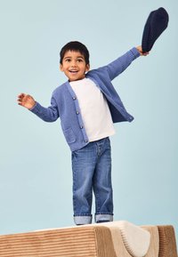 Cardigan tricotat albastru cu un buzunar, purtat peste o cămașă albă. Blugi albaștri răsuciți la manșete. Copil ținând o pălărie albastră închisă, zâmbind.
