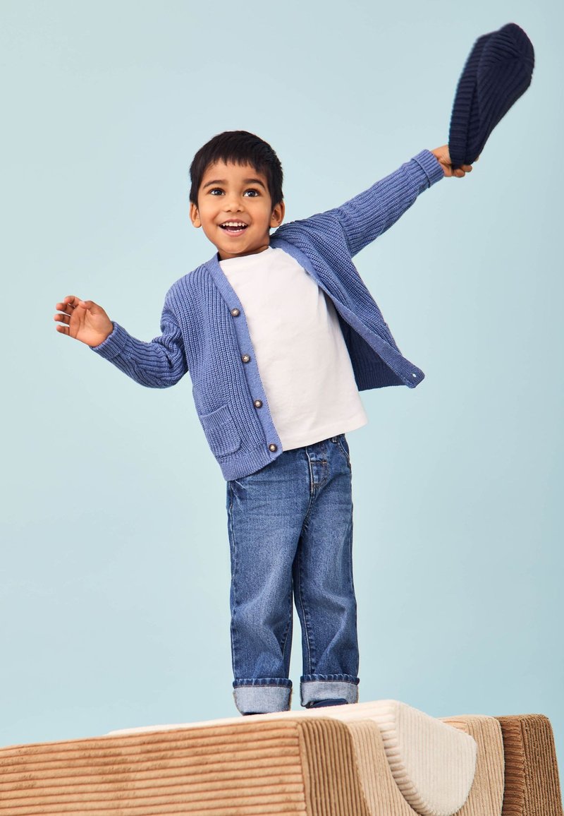 Cardigan tricotat albastru cu un buzunar, purtat peste o cămașă albă. Blugi albaștri răsuciți la manșete. Copil ținând o pălărie albastră închisă, zâmbind.