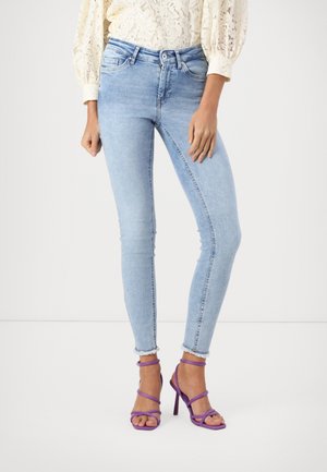 Femme portant un jean skinny bleu clair, un chemisier en dentelle crème et des sandales à talons hauts violettes avec des brides, debout sur un sol blanc.