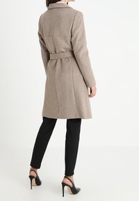 Beige wool blend coat with a belted waist, tailored design, notched collar, and back seams. Worn over black fitted pants and heels.