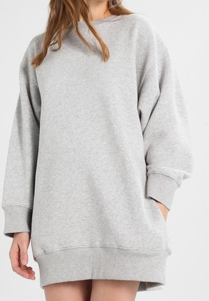 Kobieta w za dużej, jasnoszarej sukience-bluzie z prążkowanymi mankietami i dołem, stojąca z jedną ręką w kieszeni na białym tle.