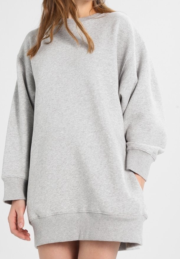 Femme portant une robe sweat-shirt ample de couleur gris clair avec des poignets et un ourlet côtelés, debout avec une main dans la poche sur un fond blanc.