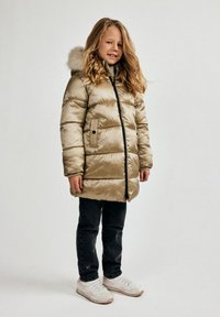 Beige pufferjakke med hætte, pelskant og frontlynlås. Har horisontal quilted design og to sidelommer, båret over mørke jeans og sneakers.