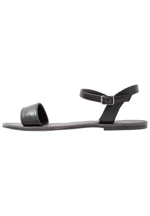 Schwarze flache Sandalen mit einem breiten Zehenriemen und einem schlanken Knöchelriemen mit einer silbernen Schnalle. Texturierte Sohle mit minimalistischem Design.