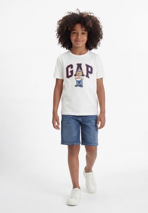 Dieťa s kučeravými vlasmi, oblečené v bielom tričku GAP s grafikou medveďa, rifľové krátke nohavice a biele tenisky, kráča dopredu na bielom pozadí.