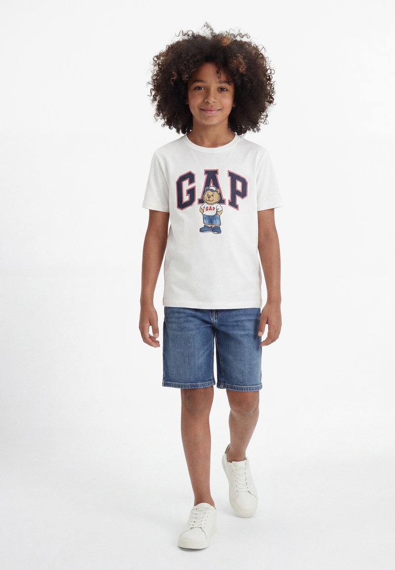 Παιδί με σγουρά μαλλιά, φορώντας λευκό t-shirt GAP με σχέδιο αρκούδας, τζιν σορτς και λευκά αθλητικά παπούτσια, περπατάει μπροστά σε λευκό φόντο.