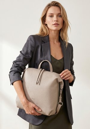 Femme aux cheveux châtain clair porte un blazer noir et une robe verte, tenant un grand sac à dos en cuir beige avec double fermeture éclair devant un fond neutre.