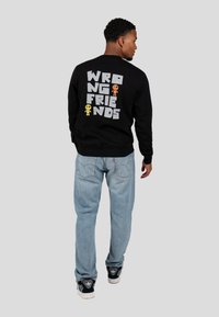 Zwarte sweatshirt met vetgedrukte witte tekst "WRONG FRIENDS" en grafische karakters, gecombineerd met lichtblauwe spijkerbroek en zwarte sneakers.