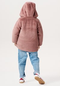 Roze fleece hoodie met een capuchon, zachte textuur en een relaxte pasvorm, gecombineerd met lichtblauwe jeans en witte schoenen met rode accenten.