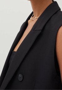 Sleeveless black blazer featuring a wide lapel, single button closure, and smooth fabric. Close-up view highlights neckline and texture.