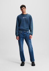 Pulover tricotat albastru cu guler clasic și logo, asociat cu blugi drepti de culoare albastru închis și pantofi negri. Design curat, fără modele vizibile.