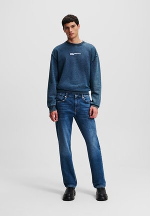 Blue knitted sweatshirt with crew neck and logo, paired with straight-leg dark blue jeans and black shoes. Clean design, no visible patterns.
