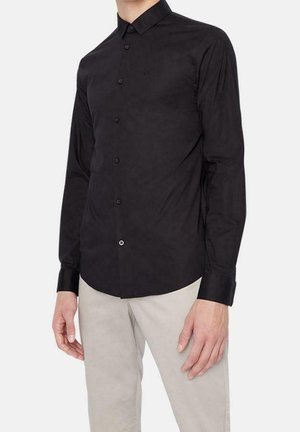 Mężczyzna w dopasowanej czarnej koszuli zapinanej na guziki z długimi rękawami i beżowych spodniach, stojący na białym tle.