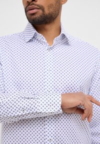 Camicia bianca con bottoni caratterizzata da un motivo geometrico a diamante blu, con colletto strutturato e polsini con bottoni. Tessuto morbido.