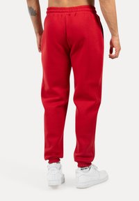Pantalones de chándal rojos con cintura elástica, bolsillos laterales y piernas ajustadas. Llevados con zapatillas blancas con detalles en negro en los calcetines.
