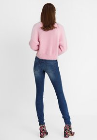 Suéter rosa de felpa con mangas anchas, combinado con jeans skinny de color azul oscuro. El conjunto incluye zapatos de tacón alto con acentos florales.