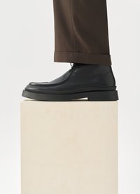 Svarta läderskor med rundad tå, fästa vid en kraftig sula, med synlig sömnad. Matchade med brun skräddarsydd byxa.