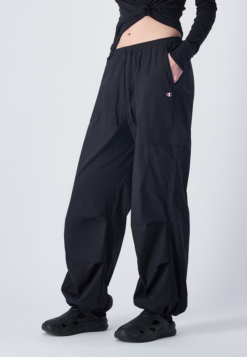 Pantaloni cargo neri larghi con cintura elastica, tasche laterali e scarpe nere senza lacci, indossati da una persona in piedi contro uno sfondo semplice.
