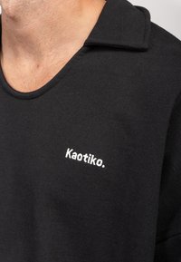 Schwarzer Sweatshirt mit rundem Halsausschnitt und kurzen Ärmeln. Auf der linken Seite befindet sich gestickter, weißer Text "Kaotiko." Weiche Textur.