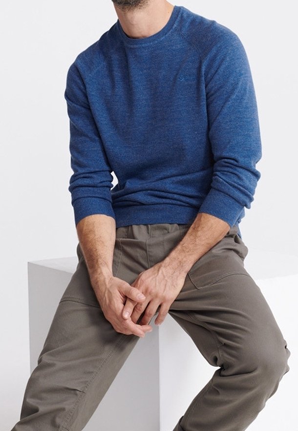 Homme portant un pull bleu à manches longues et un pantalon gris, assis sur un cube blanc avec les mains légèrement jointes dans le giron.