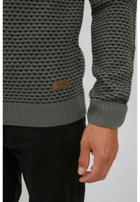 Suéter de punto gris oscuro con un patrón texturizado, puños y dobladillo acanalados, y un parche de logo marrón en el lado inferior.